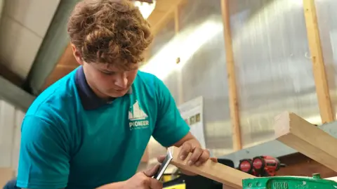 Martin Giles/BBC Lewis has curly brown hair, and he is working at his workbench. He has a piece of wood in one hand, and a chisel in the other. He is wearing a turquoise-coloured T-shirt.