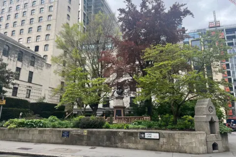St Mary Aldermanbury park with a small grey brick wall surrounding a raised park with trees, plants and benches inside it