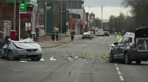 BBC A grey-blue Ford Focus lies across a road with its back smashed and much of its roof sliced off. A dark-coloured police car, with its front damaged and back gate open, stands opposite. Debris is scattered across the cordoned-off section of road. Yellow crash-scene investigation markers have been put in place on the road.