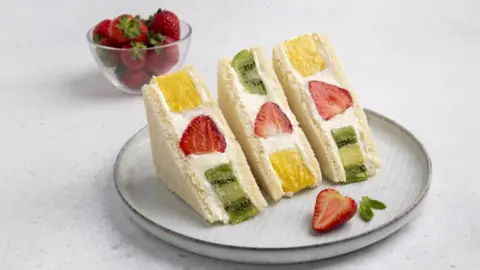 Dariia Chernenko/Getty Images Three sandwiches in white bread on a plate. Each sandwich contains cream, a strawberry, a piece of kiwi and a piece of pineapple. In the background is a small bowl of strawberries
