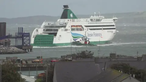 An Irish Ferries ship sits close to the port with a road and some infrastructure seen in the foreground. There is a support boat by the side of the ferry helping it to dock.