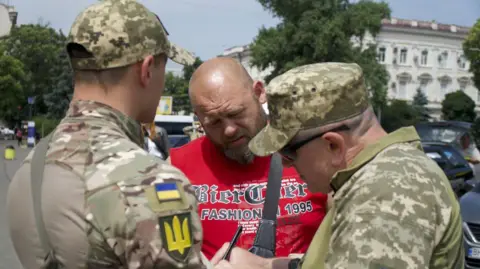 Thanyarat Doksone/BBC Conscription officers Anatoliy (in sunglasses) and Oleksiy stop a man on the streets of Odesa 