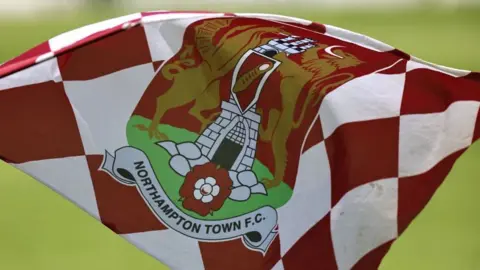 A white and red chequered flag with a Northampton Town F.C. badge in the middle of it. Behind the flag is grass. 