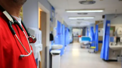 A ward in a hospital with a number of blue curtains on either side of a corridor in front of beds. A staff member on our left has a red top and a stethoscope around their neck.