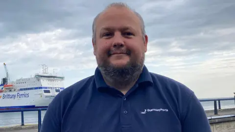 Joe Armstrong from Guernsey Ports is stood beside the bridge looking straight at the camera and is smiling. He is wearing a navy polo shirt and there it a large Brittany Ferry in the background. 