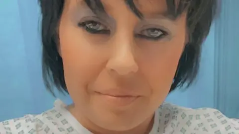 Cancer Research UK Rebecca Dance is pictured on a hospital ward and is wearing a patient gown. She has dark, short hair and is wearing make-up. 