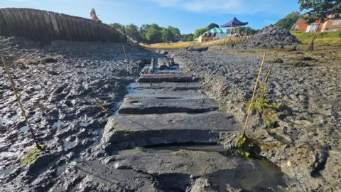 Buckler's Hard Shipyard Trust and Maritime Archaeology Trust Image shows the exposed slipway. The image is taken down low close to the exposed groundways. Either side of the beams is mud. At the end of the slipway, is a green and white tent which is being used to show the artefacts found and explanations of the digging taking place.