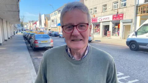 Clive Burns standing on the edge of a pavement. Photographed from waist up. He is wearing red glasses and a grey/green jumper.