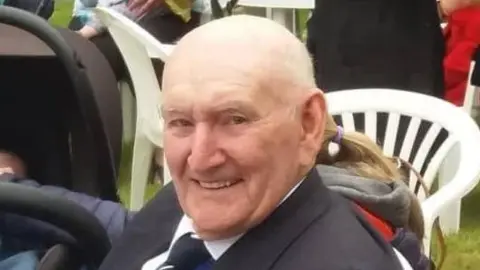 PHIL GAWNE Raymond Gawne has a bright smile and wears a suit as he sits outside at a family event.