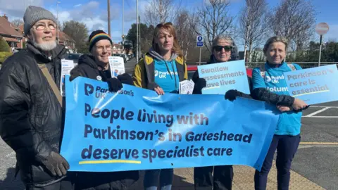 A group of people wearing winter outwear and blue t-shirts reading Parkinson's UK stand together. They are standing on a street holding a blue banner which reads - people living with Parkinson's in Gateshead deserve specialist care.