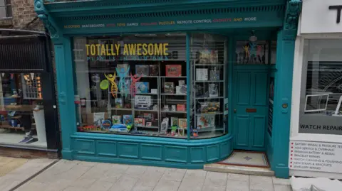 Google A toy shop with a turquoise door and window frame in a street in York. The words Totally Awesome are in the window and there are lots of toys.