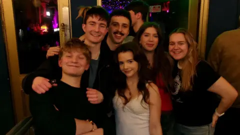 Freya Price Six people stand closely together for a photo at the entrance of a music venue. The group is framed by gold-coloured doors and windows, with a bright green “FREE ENTRY” sign overhead and colourful lights visible inside.