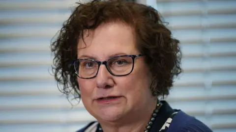Dr Hilary Cass speaking to reporters in 2024. She has short, dark, curly hair and dark-framed glasses. She is wearing a blue cardigan over a black and white patterned top, with a navy beaded necklace. 
