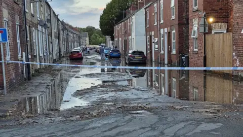 Police tape cordening off a partially flooded road with terraced houses on each side along with a number of cars.
