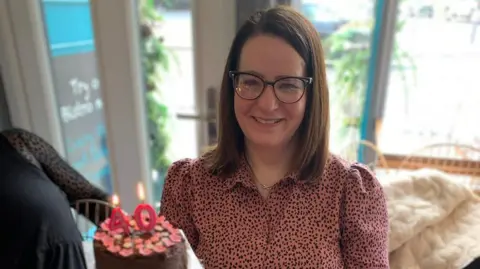 Anona Gauld is wearing a pink and black spotty shirt and black glasses. She is smiling and there is a 40th birthday cake in front of her. 