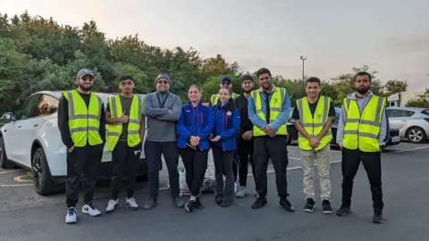 iCare Peterborough People standing in a row- wearing high-visibility green jackets- and Akeel (3rd from left) wearing a grey jumper and blue jeans