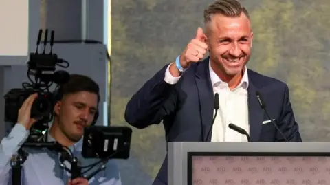 Ulrich Siegmund, who has a dark blonde undercut which is swept back on the top, grins at his audience from a lectern and does a thumbs up 