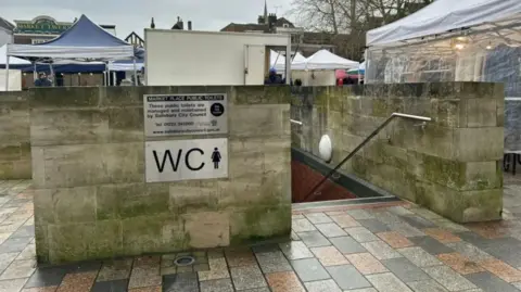 The previous underground toilet block. It shows a concrete wall and staircase leading below ground. It is positioned in the middle of a market square with stalls and marquees surrounding it. 