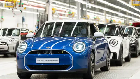 A row of shiny new Mini Cooper Electrics on the production line at the BMW Mini plant. The first is blue, the second white, the third black.