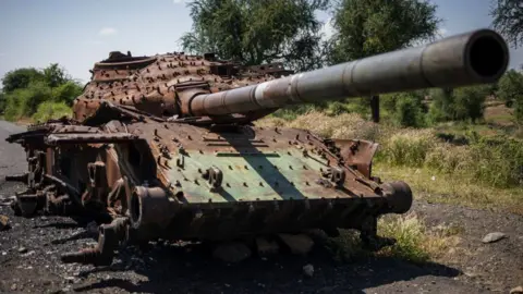 A rusted, wrecked tank lies on the side of a rural road. Mountains are visible in the background.