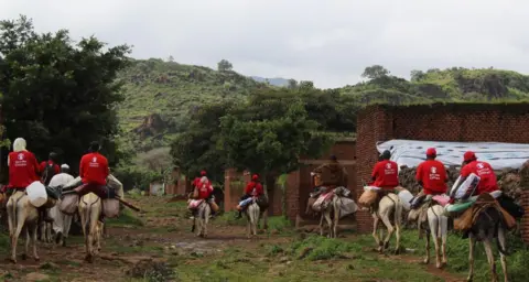 Save the Children Aid workers in red T-shirts riding on donkeys carrying luggages down a hilly terrain  