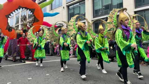 Six young people marching in a parade. They are wearing black trousers, a green costume and gold helmets. There is an inflatable orange ring being pulled along behind them. 