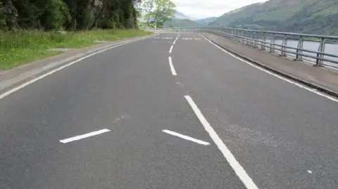 Transport Scotland A white arrow-like marking on a road before a bend. On one side of the road is a large loch with hills on the opposite shore.