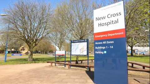 Outside entrance of New Cross Hospital, trees in the back ground with a sign with NEW CROSS HOSPITAL on it with writing saying emergency department below