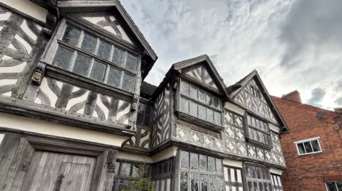 A black and white, three-storey Elizabethan building. There is a brick building to the side of it. A wooden door in the lower left corner of the image has a smaller door within it. There are ornate features - including animal carvings - in the wooden structure of the building. Panelled windows take up large portions of the building's frontage.