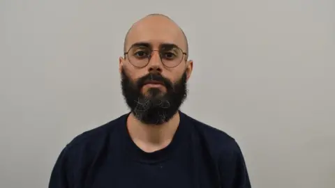 Metropolitan Police Police mugshot of Abdullah Albadri. He has very short hair and a longer beard with a few grey hairs. He wears a dark coloured jumper and stands in front of a dark grey background. 