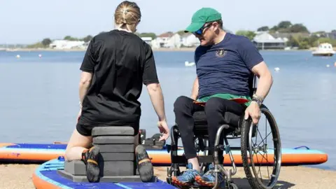 Amy Tallula Will Behenna sat in a wheelchair with a green cap and sunglasses on working with a woman who is kneeling on a paddleboard with blocks behind her assisting her