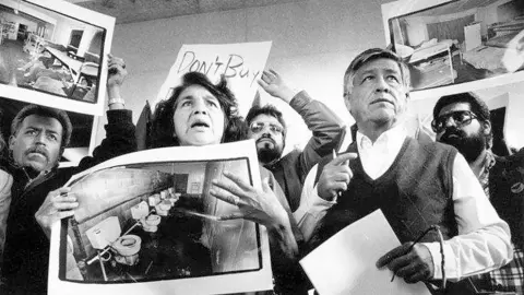 Getty Images Dolores Huerta holds a poster-sized image of toilets while standing next to Cesar Chavez. The image is a black and white image from when the pair was younger. Other people crowd behind thme in the image. 