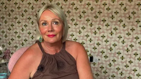 Jo has blonde hair and is wearing a brown dress. She is sitting behind some wallpaper. 