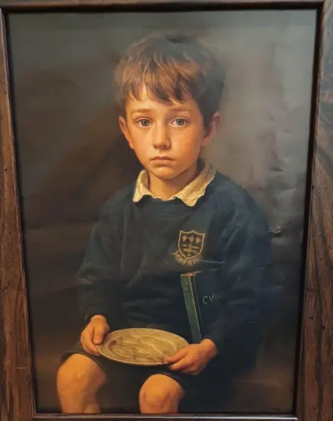A boy sitting on a chair in school uniform holding an empty plate - a digital print. Inside a wooden frame