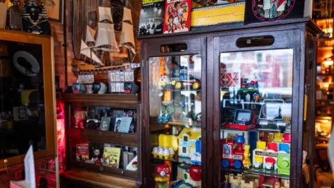 Jack Merriman A cabinet full of toys and trinkets which are used during the piano lessons. The room is packed with old-fashioned toys and mismatched items.