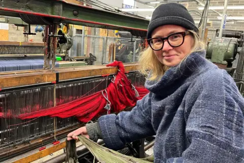 Clare is working at a loom at her business in Evanton. She is wearing a thick woolly coat and a hat.