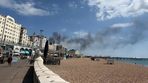 Smoke is seen over a beach. In the background are white Regency buildings and a Victorian pier. 