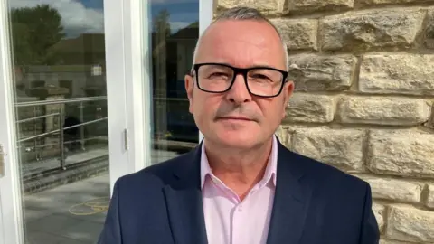 Lee Barron with short white hair and large black glasses looking at the camera wearing a dark blue jacket and pink shirt in front a stone wall with a patio-style door