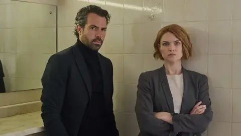 Tom Jackson/Severn -scherm Tom Cullen en Erin Richards stonden in een badkamer, met witte tegels en spiegels erachter. Tom, aan de linkerkant, draagt een zwart pak en zwarte polosnek top en heeft krullend donker haar en een baard. Erin heeft aan de rechterkant kort gekrulde gemberhaar en draagt een grijs pak en een witte top. Ze kijken allebei naar de camera met serieuze uitdrukkingen.