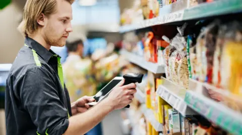 Getty Images Young male supermarket worker, dressed in a dark shirt with green trimming, scans food on a shelf 