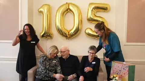 Richard Knights/BBC Ron sits on a sofa animatedly chatting to a woman on his right. The women around him, some of whom are care home workers, are all laughing. There is a gold 105 balloon behind them.