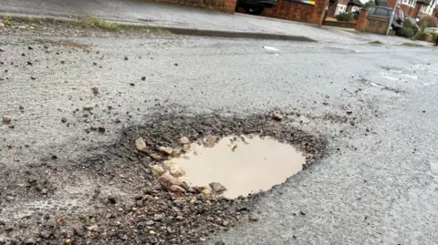 A deep pothole on a residential street, filled with rain water. Walls at the edges of resident's drives can just be seen beyond.