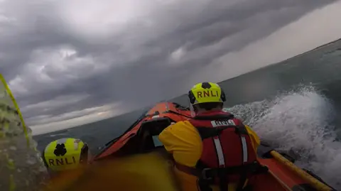 A view from behind of two RNLI crew members. They are on a lifeboat with waves splashing about. The sky above them is dramatic - dark and cloudy.