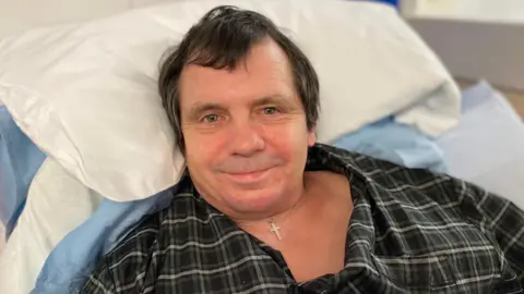 Spencer Wood is wearing a black and white plaid shirt and has brown hair. He is lying on a bed with pillows behind his head. 