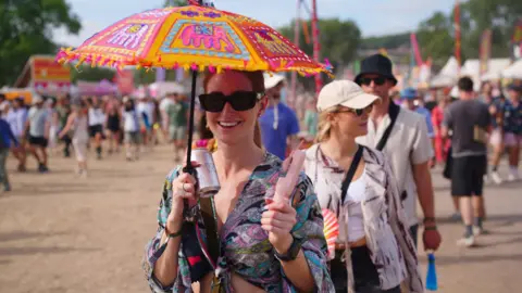 PA Media A woman is holding a colourful umbrella to shield from the sun. In her other hand she is holding a portable fan. She is in a crowd of people and the sun is shining.