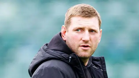 Finn Russell standing on the pitch wearing a black coat looking over his right shoulder before Bath v Leicester.
