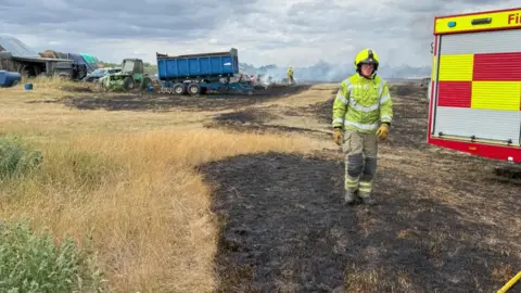 Essex County Fire and Rescue Service A firefighter stands on a scorched field beside a fire engine, behind him is a blue tipper truck and other firefighters. Smoke is rising from the horizon.