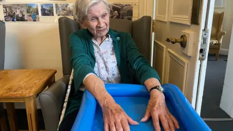 A woman sits in a chair indoors with her hands in a blue paddling pool, with photos and furniture around her.