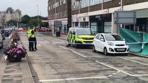 Police tape cordoning off road in the daylight. There are two police officers stood in the road and police vehicle parked alongside a white car.