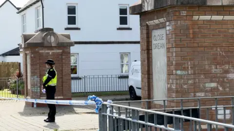 A policewoman stands at the entrance of Close Drean behind police tape cordoning it off.